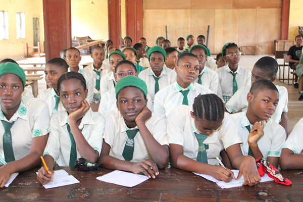 Students in white uniforms and green ties sit behind a long desk awaiting class to begin.