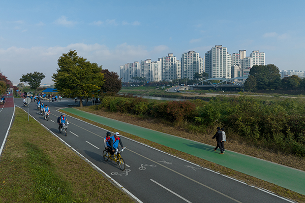 A row of bicycles progresses down a bike path near a green walking trail, skyline of the city in background.