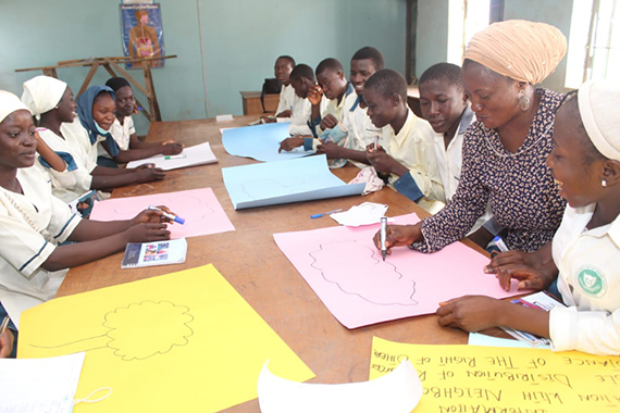 A women draws a cloud on a pink sheet of paper while several other people around a table watch