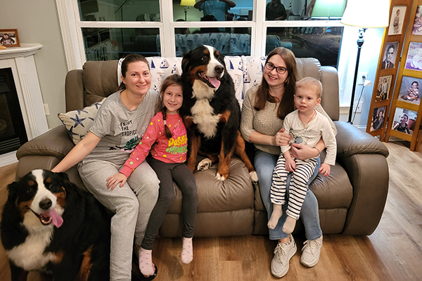 The two women sit on a sofa with their children in their laps, and two dogs nearby