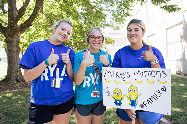 Three young women give the thumbs up while holding a sign that says Mike's Minions