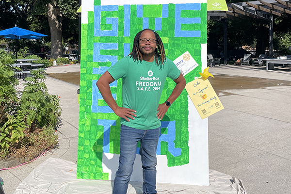 Bespectacled young man in a green ShelterBox shirt stands in front of a handmade sign